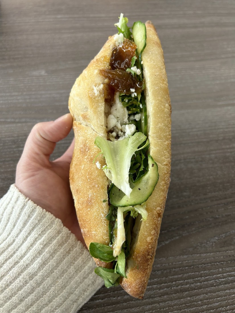An open sourdough sandwich filled with soft cheese, chutney, cucumber and salad leaves, held in hand